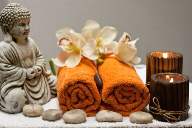 Mindfulness-inspired spa setting with a Buddha statue, lit candles, and rolled orange towels, symbolising relaxation, self-care, and emotional wellbeing.